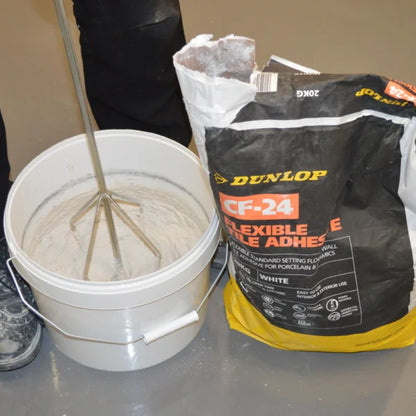 Bucket of mixed adhesive with a Dunlop CF-24 flexible tile adhesive bag on a gray surface.